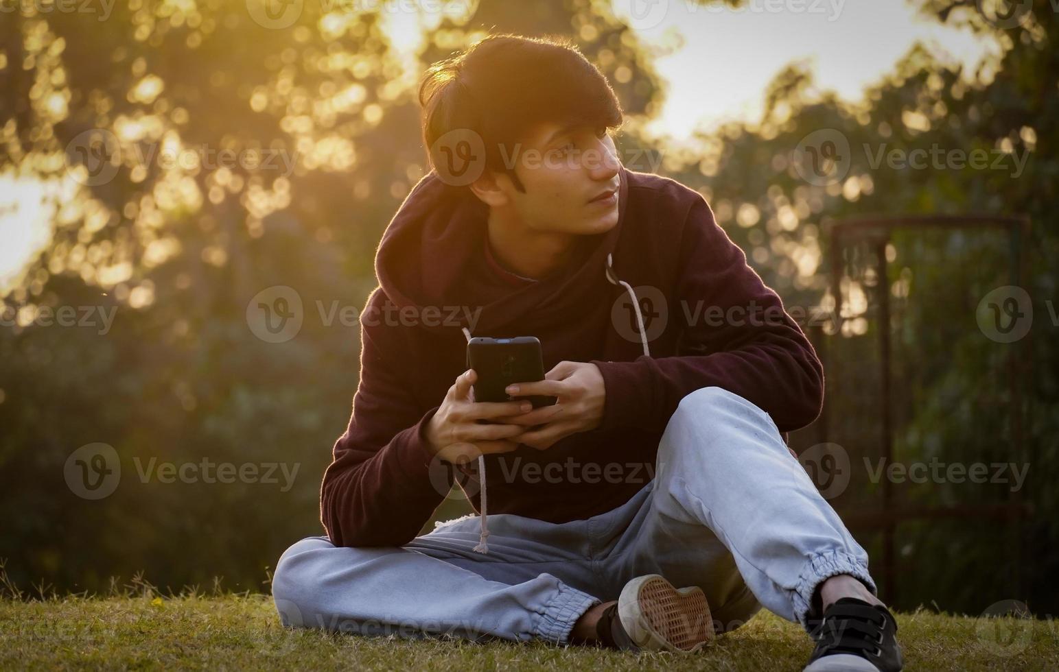 A male student using phone seated on a grass in the city park photo