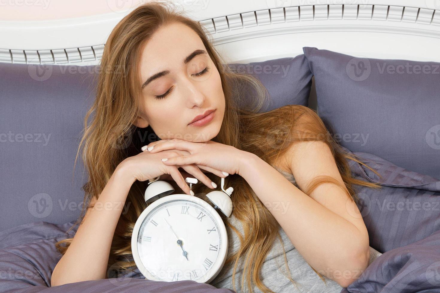 Concept of overslept work, bad sleep insomnia - Young beautiful sleepy girl with a closed eyes holds a clock and lies in her bed in the bedroom in the morning photo