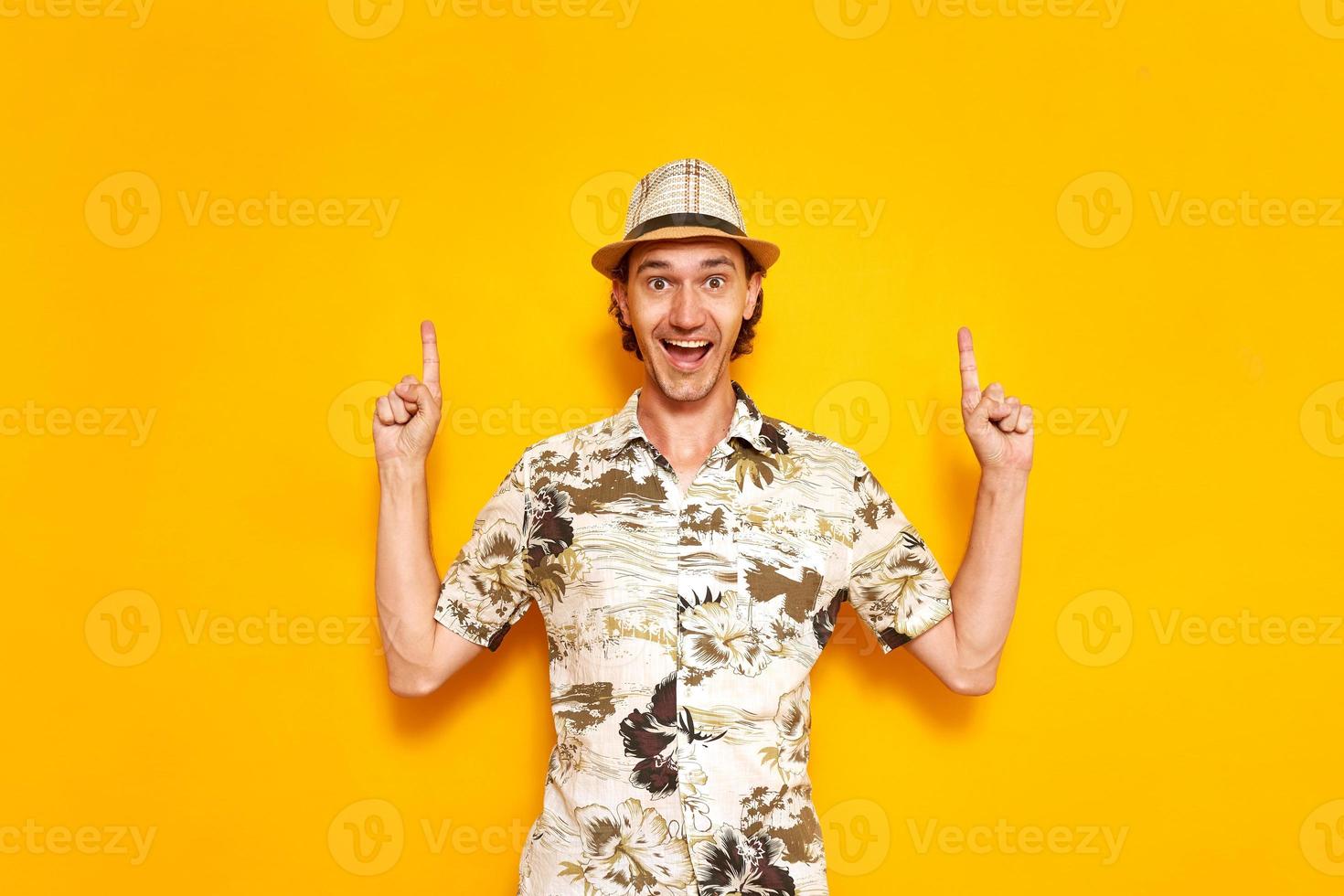 a young happy male tourist isolated on a yellow background points his index fingers up at an empty area. He is wearing a hat, a Hawaiian shirt. space for text. Concept - recreation, tourism, people. photo