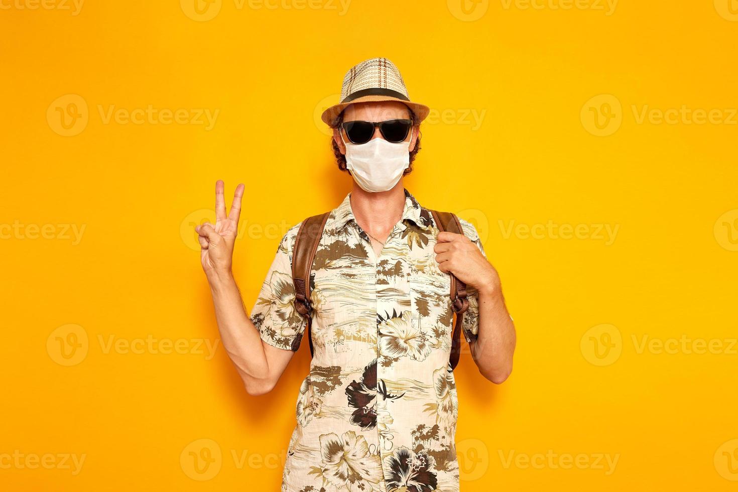 young male tourist traveler with backpack in a hat, sunglasses, a medical mask shows a peace sign with his hand isolated on a yellow background. the concept is recreation, people, adventure, vacation. photo
