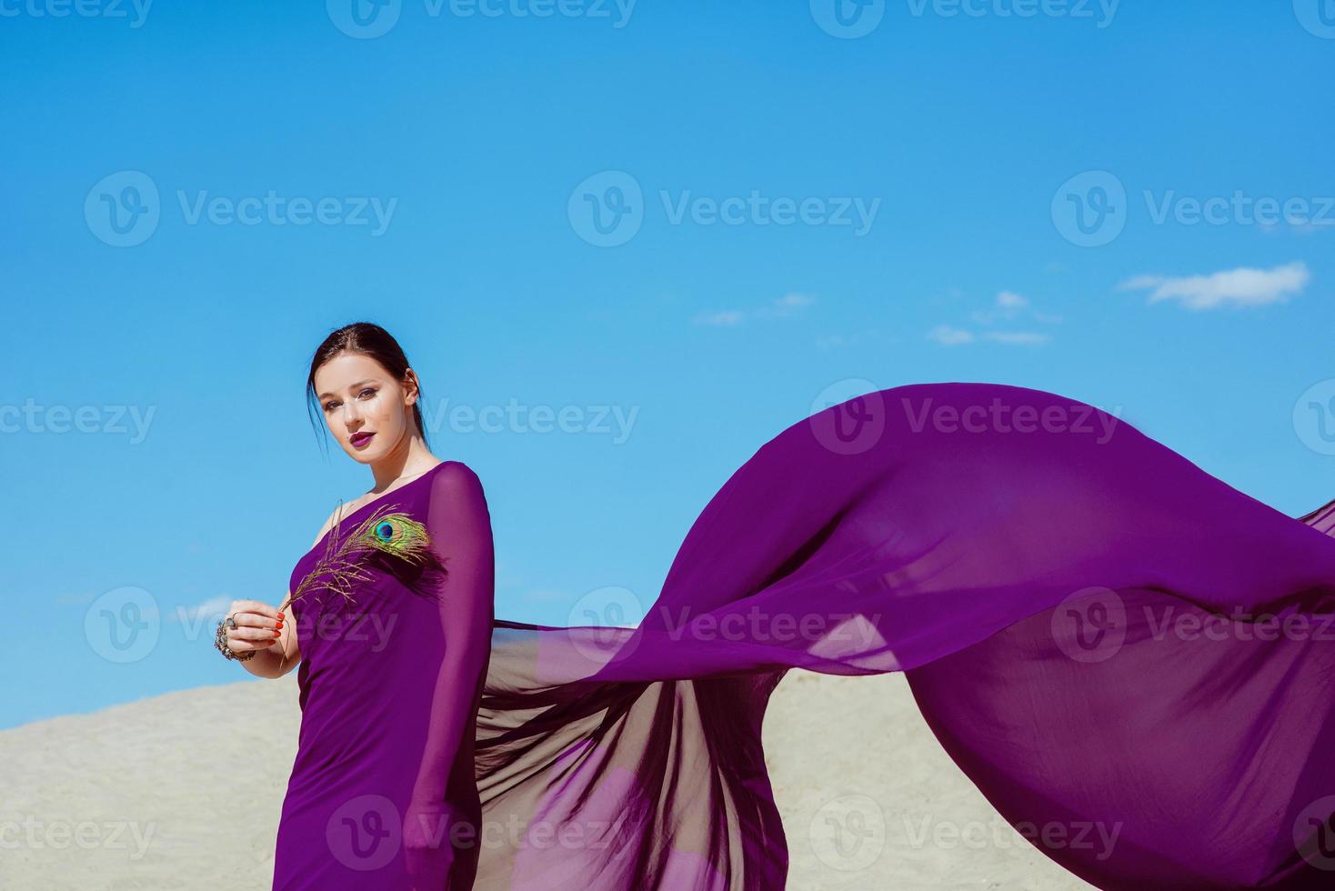 increíble hermosa mujer morena con la pluma de pavo real en tela púrpura en el desierto. oriental, indio, moda, concepto de estilo foto