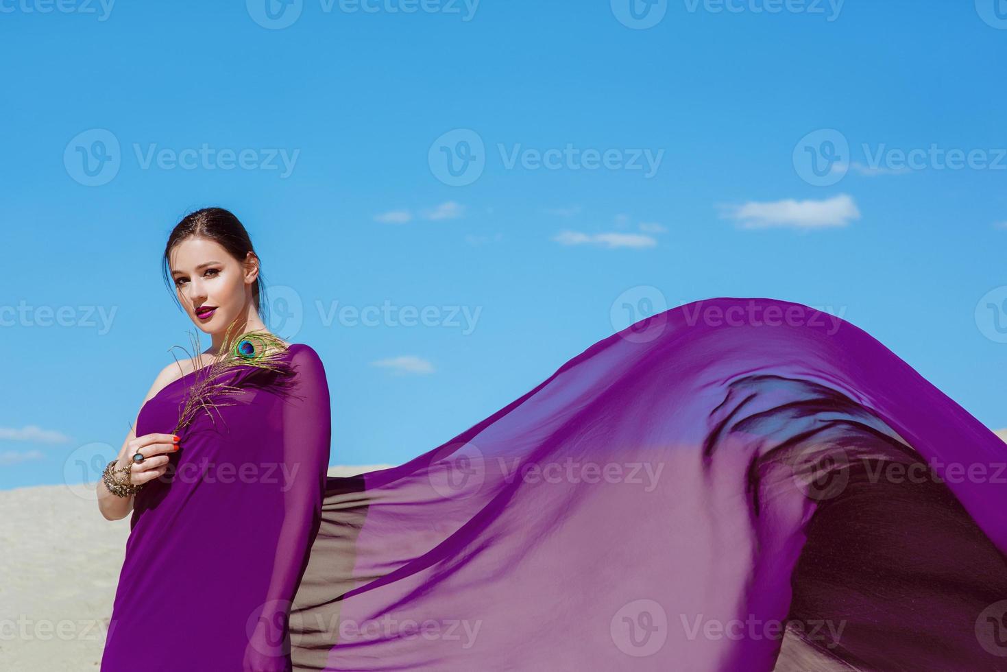 increíble hermosa mujer morena con la pluma de pavo real en tela púrpura en el desierto. oriental, indio, moda, concepto de estilo foto