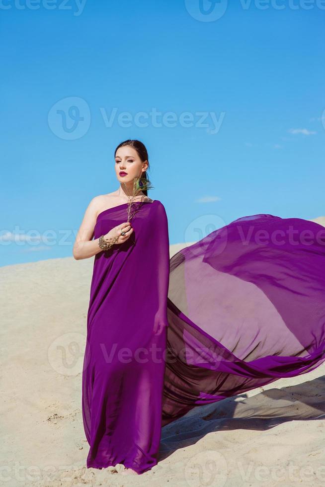 increíble hermosa mujer morena con la pluma de pavo real en tela púrpura en el desierto. oriental, indio, moda, concepto de estilo foto