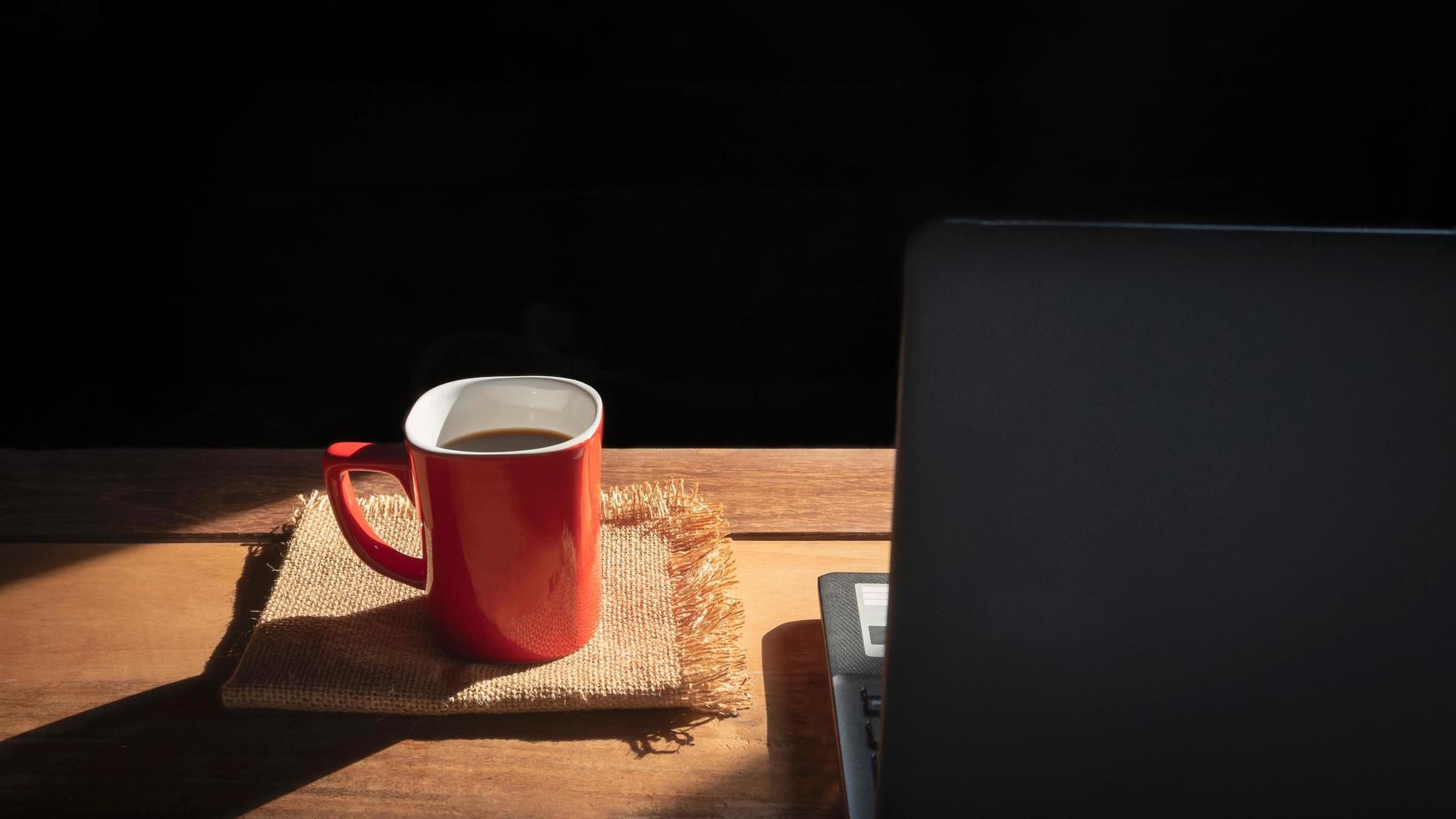 concéntrese en la taza de café roja sobre las servilletas y el portátil negro borroso sobre una mesa de madera con luz solar y sombra en el fondo negro, concepto de pausa para el café foto