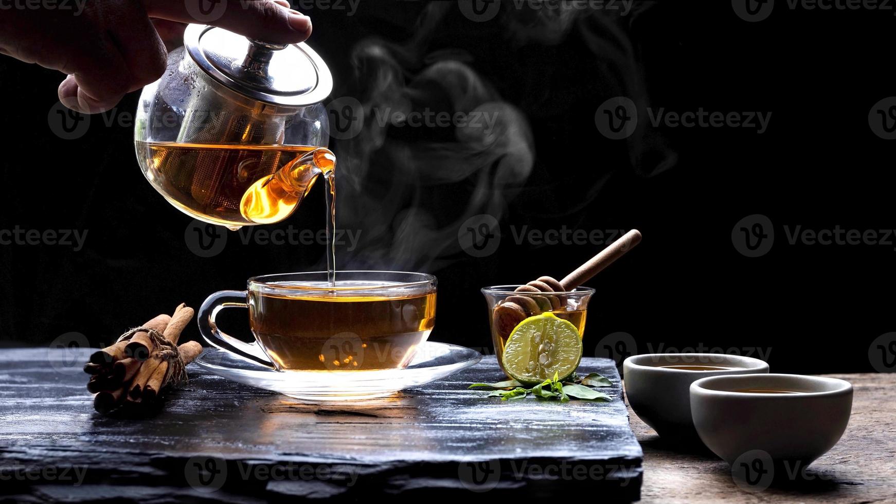 Pouring hot aromatic herbal tea from teapot into glass teacup set with steam and various herbs on black stone plate with wooden table floor in dark background, selective focus photo