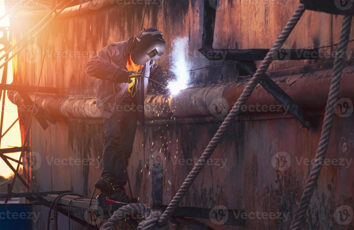 la vista lateral del soldador en ropa de trabajo protectora en la plataforma está soldando el viejo casco oxidado del buque en el área del astillero al atardecer foto