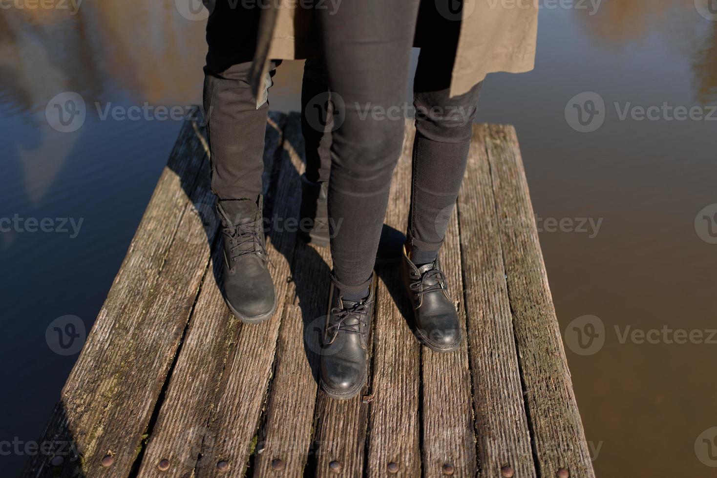 Cropped photo young legs in sneakers, married couple, husband and wife on a wooden bridge near lake. Rear view of couple standing on pier. lower half. Place for text and design. Summer in love.