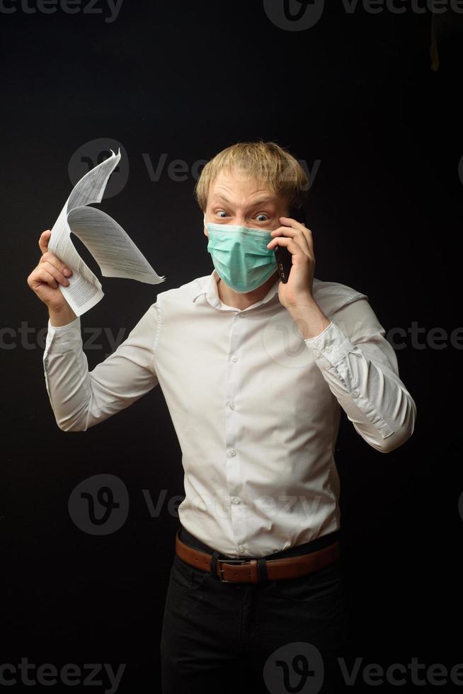 A young man in a medical mask quarrels on the phone analyzing the documents received. The concept of finding a solution to business problems due to the epidemic of coranavirus photo