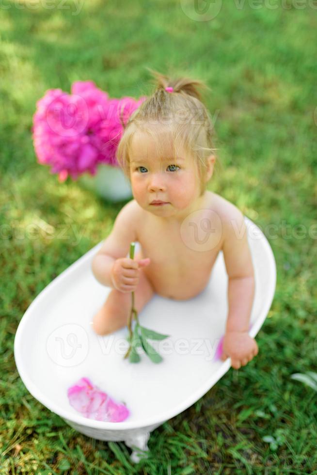 Happy toddler girl takes a milk bath with petals. Little girl in a milk bath on a green background. Bouquets of pink peonies. Baby bathing. Hygiene and care for young children. photo