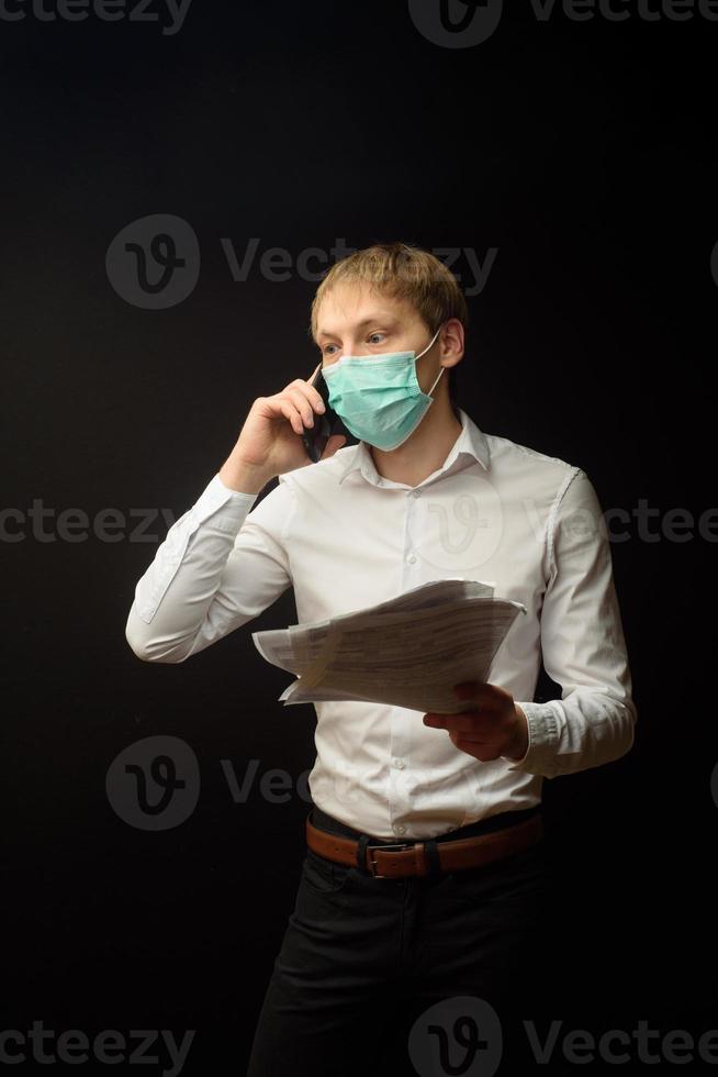 un joven con una máscara médica pelea por teléfono analizando los documentos recibidos. el concepto de encontrar una solución a los problemas comerciales debido a la epidemia de coranavirus foto