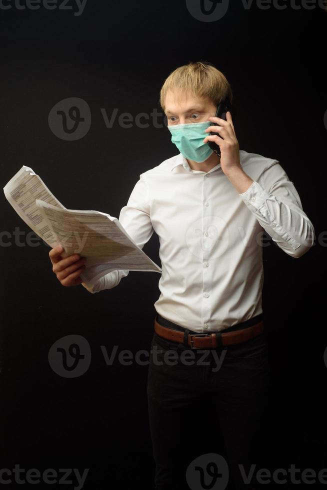 A young man in a medical mask quarrels on the phone analyzing the documents received. The concept of finding a solution to business problems due to the epidemic of coranavirus photo