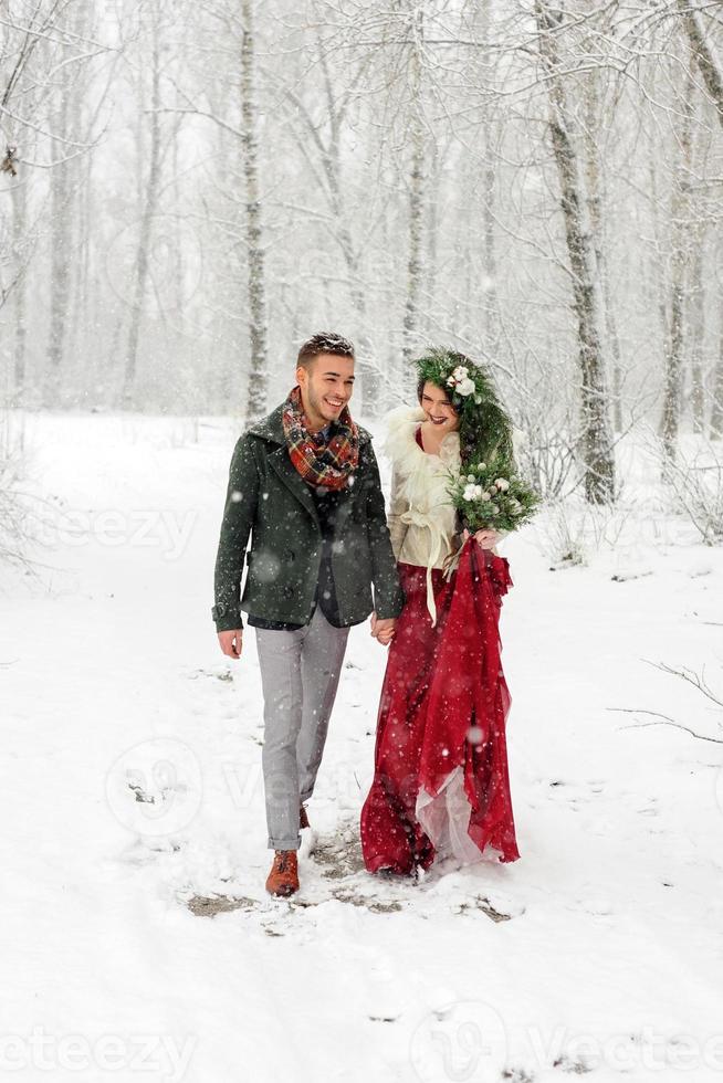 hermosa novia y novio con un perro blanco están parados en el fondo de un bosque nevado. foto