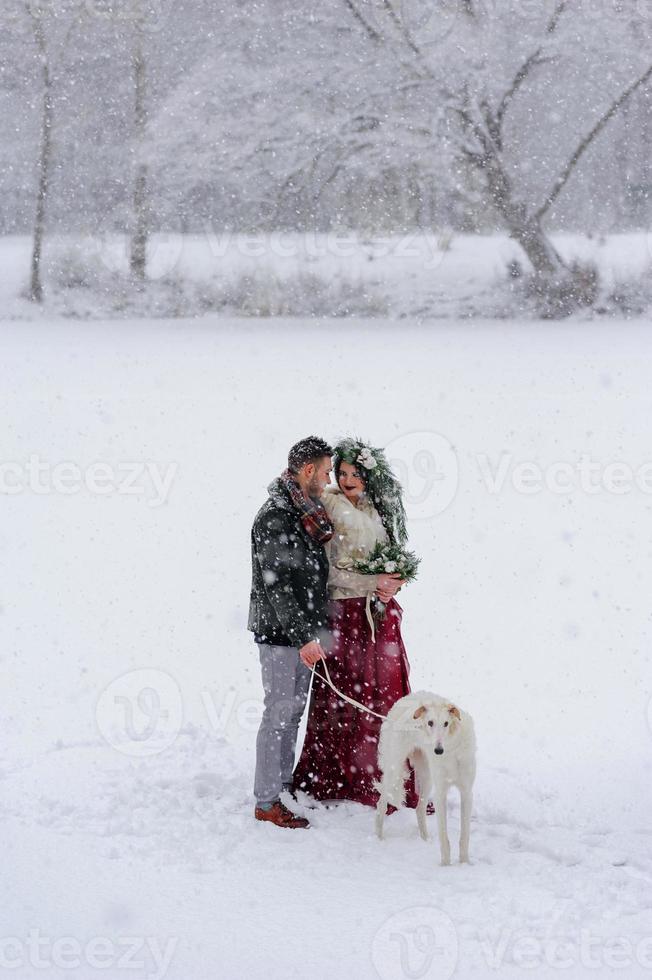 Beautiful bride and groom with a white dog are standing on the background of a snowy forest. photo