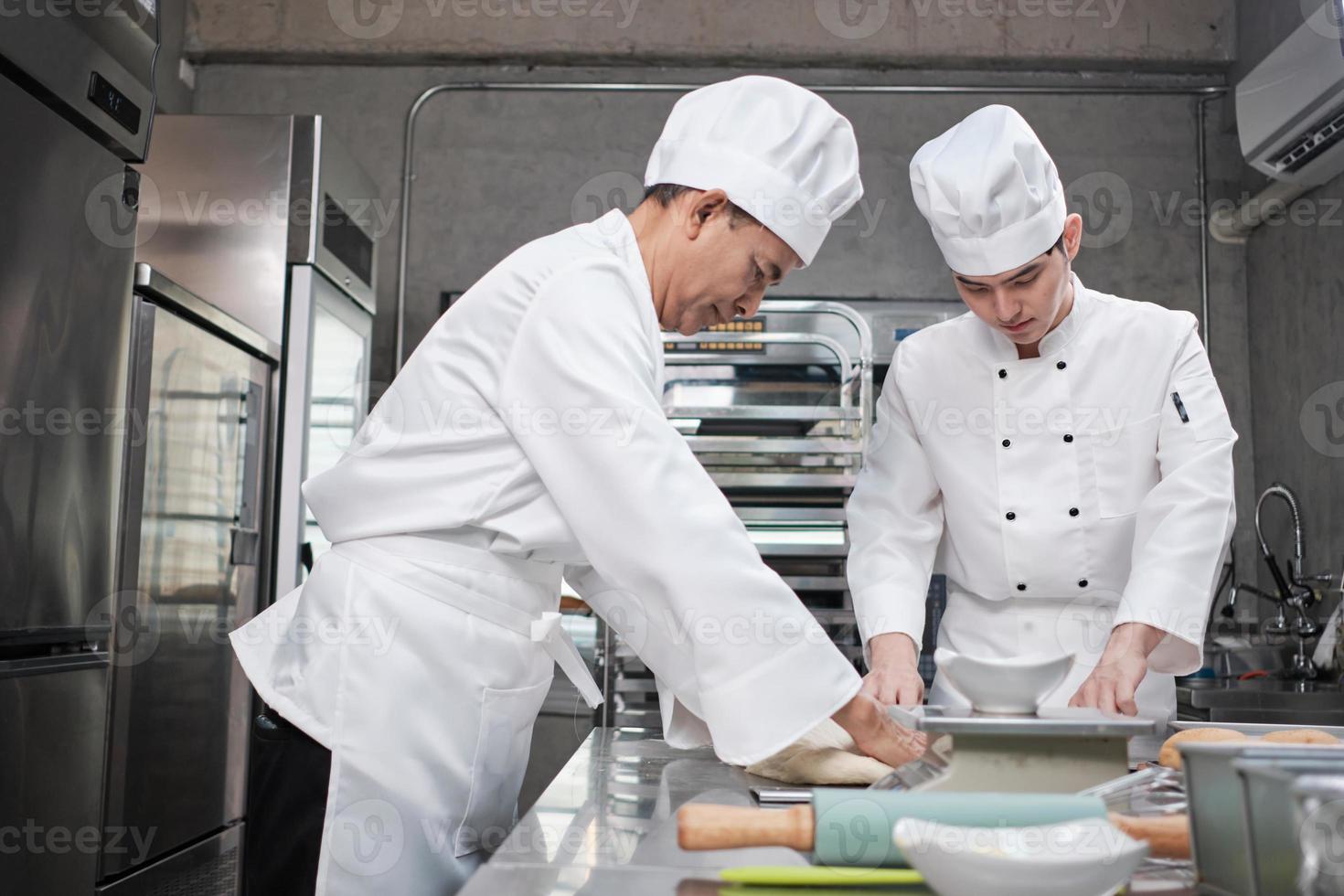 Two professional Asian male chefs in white cook uniforms and aprons are kneading pastry dough and eggs, preparing bread and fresh bakery food, baking in oven at stainless steel kitchen of restaurant. photo