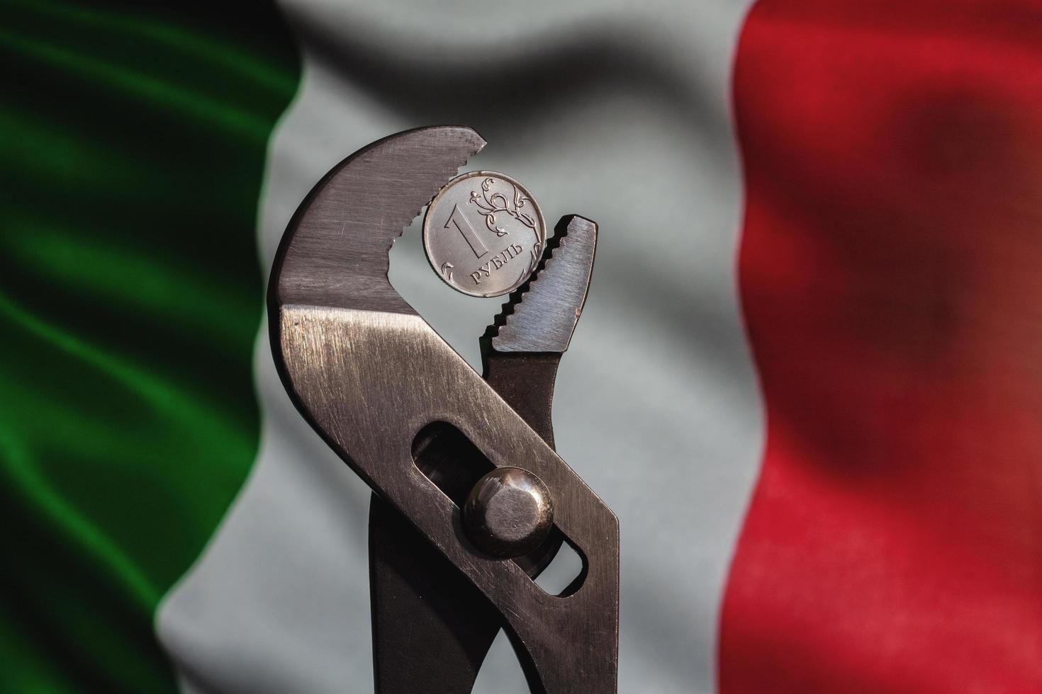 Coin one ruble is clamped in a wrench against the background of the flag of Italia. The idea of the collapse of the ruble and economy sanctions against Russia. photo