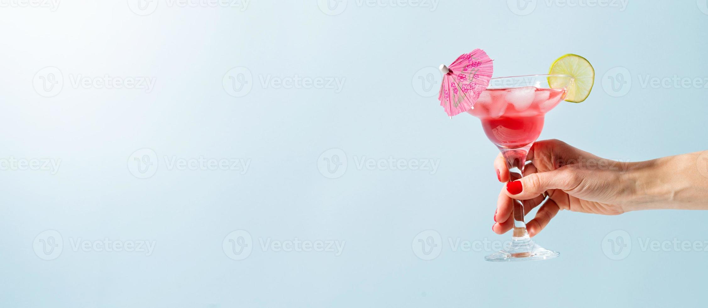 female hand with nice red manicure holding fresh summer cocktail with strawberry, lime and ice cubes on blue background with copy space photo