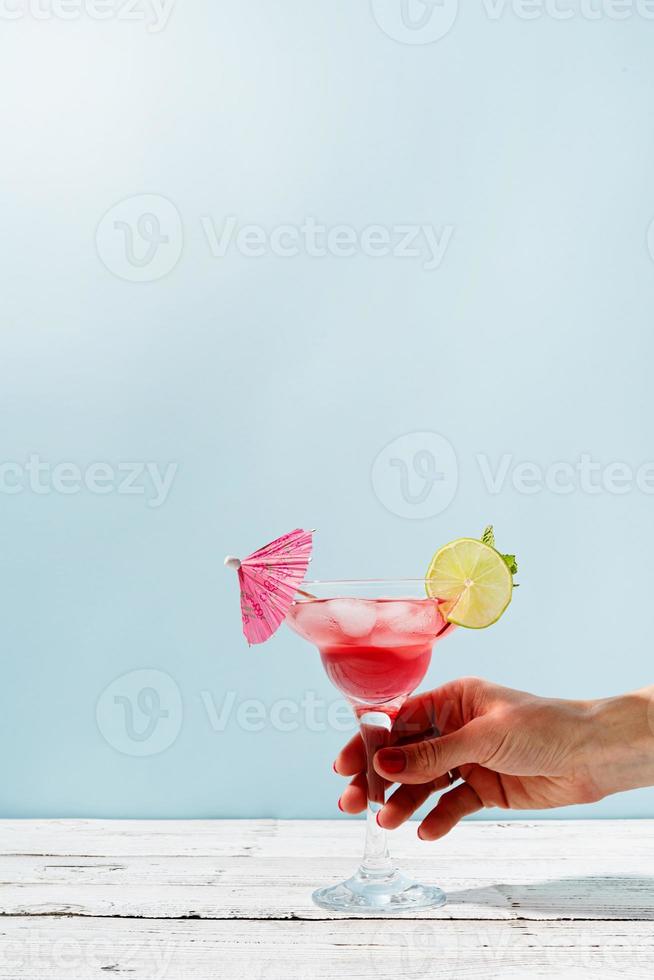 female hand with nice red manicure holding fresh summer cocktail with strawberry, lime and ice cubes on blue background with copy space photo