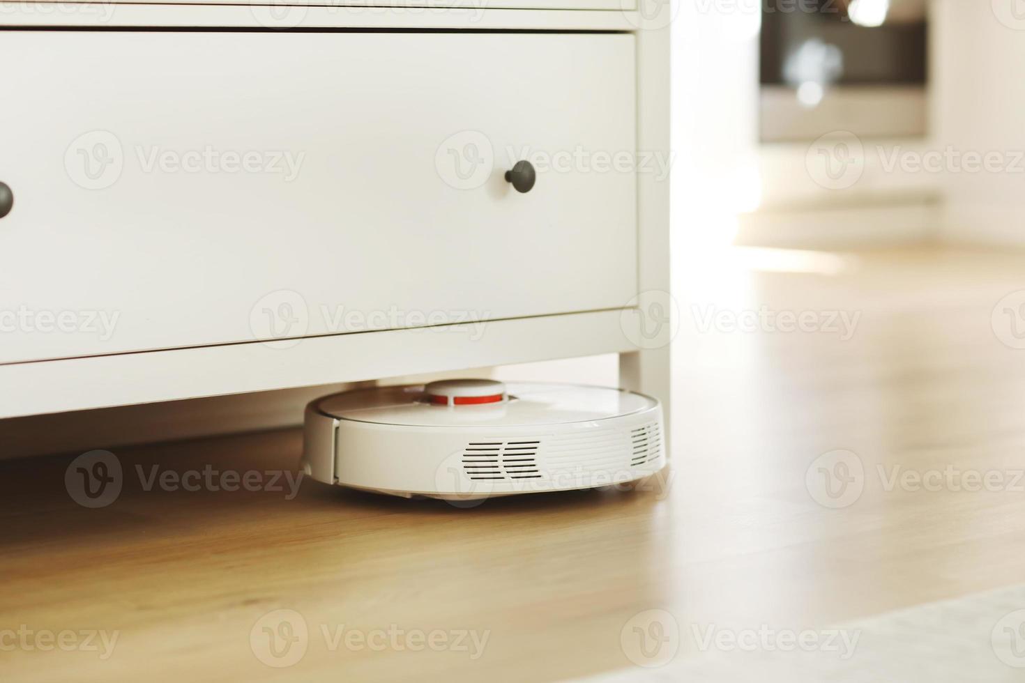 aspiradora robótica blanca en el suelo laminado limpiando el polvo en el interior de la sala de estar. tecnología de limpieza electrónica inteligente. casa inteligente foto