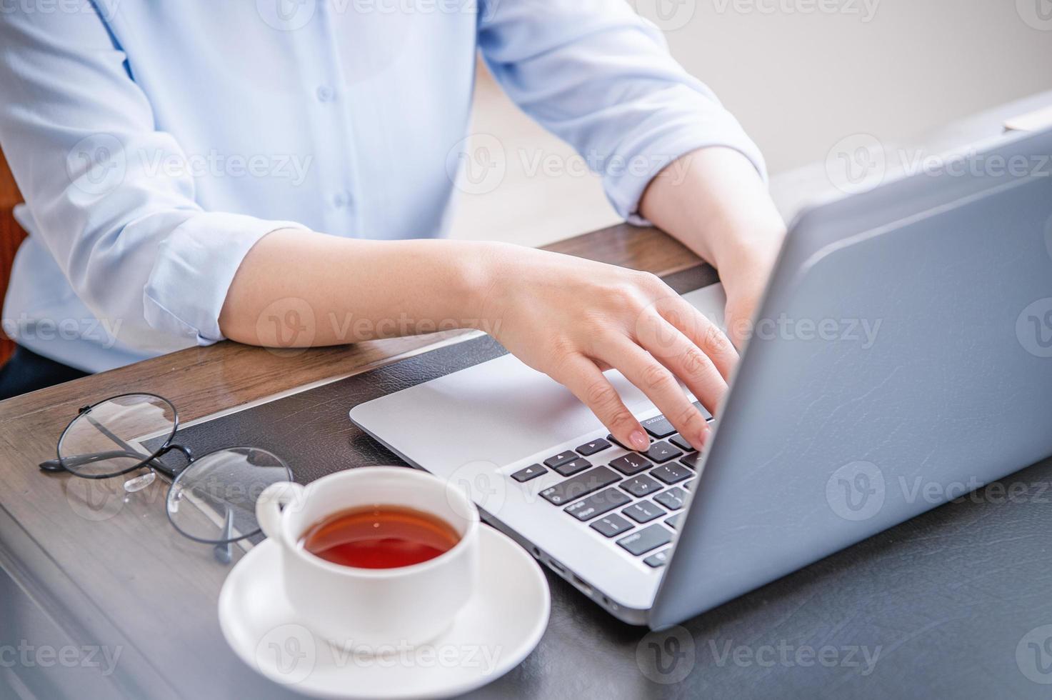 Business concept. Woman in blue shirt typing on computer with coffee on office table, backlighting, sun glare effect, close up, side view, copy space photo