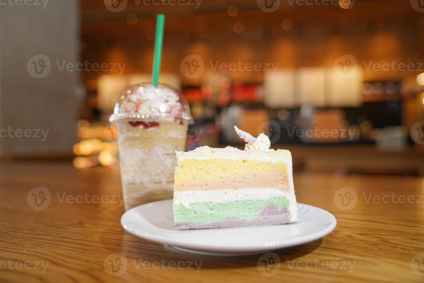 Slided rainbow cake decorate with unicorn horn on top next to coffee frappe. The cake is in a white disk on the wooden table and blur background. photo