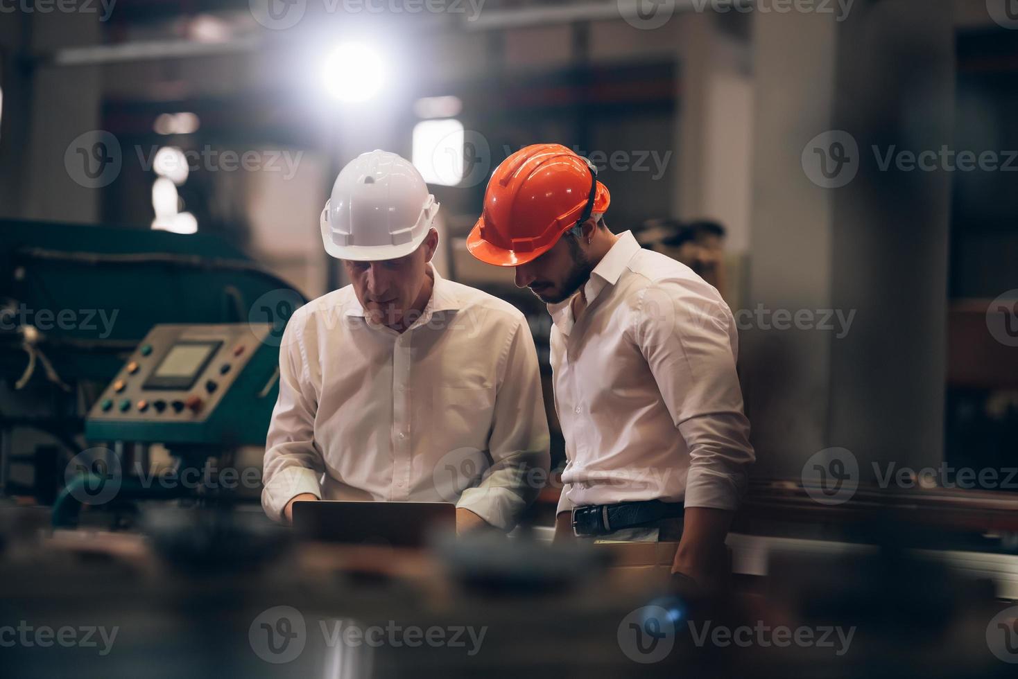 Factory worker man and engineer manager working together a metal sheet industrial worksite , checking machine process on laptop and wearing hard hat for safety photo