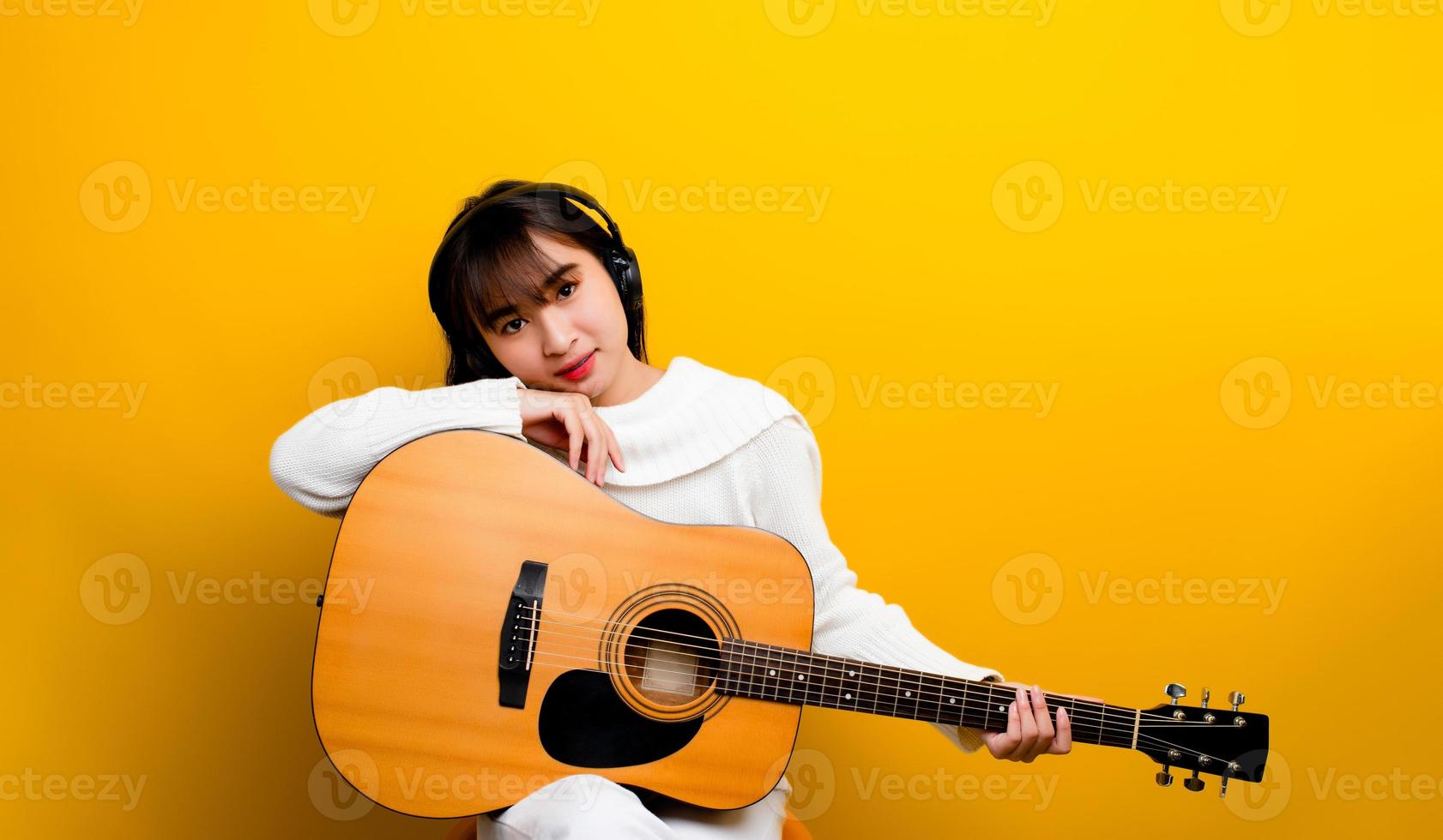 hermosa guitarrista mujer étnica asiática cantando y tocando la guitarra mientras se sienta en una silla con las piernas cruzadas. música de fondo en color y conceptos de relajación foto