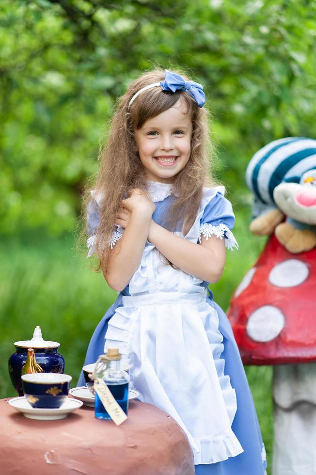 una niña linda con el disfraz de alicia del país de las maravillas organiza una fiesta de té en su mesa mágica. fotografiado en la naturaleza. foto