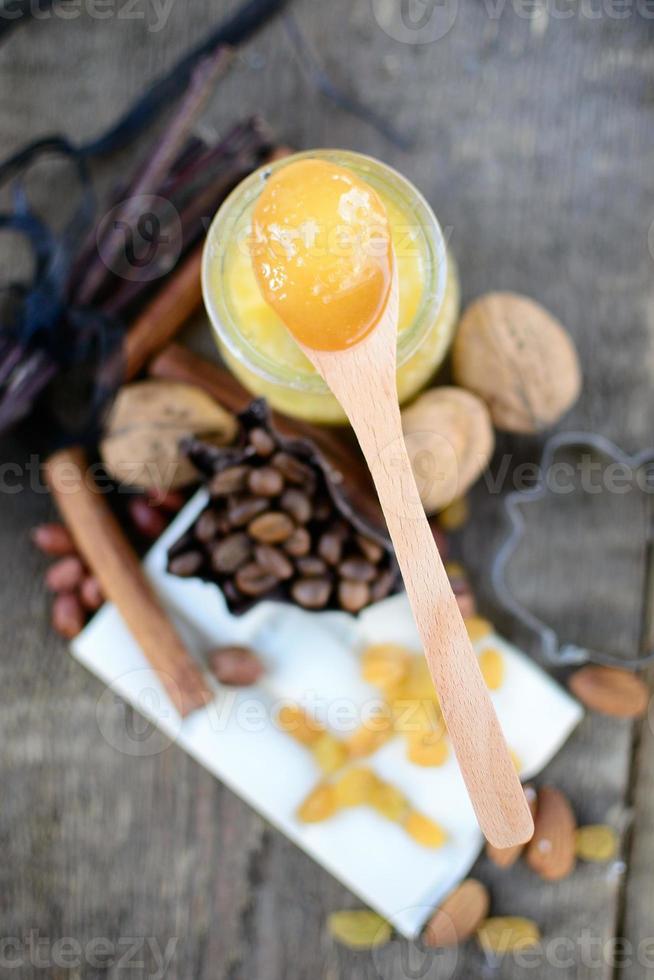 cuchara de madera con miel sobre una mesa de madera junto a baldosas de chocolate blanco, pasas, palitos de canela foto