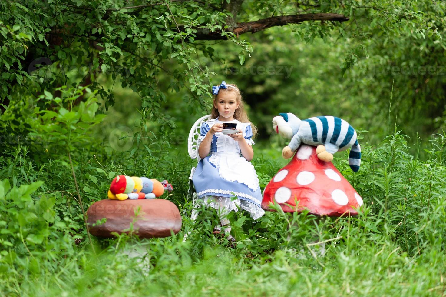 A little cute girl in the costume Alice from Wonderland holds a tea party at her magic table. Photographed in nature. photo