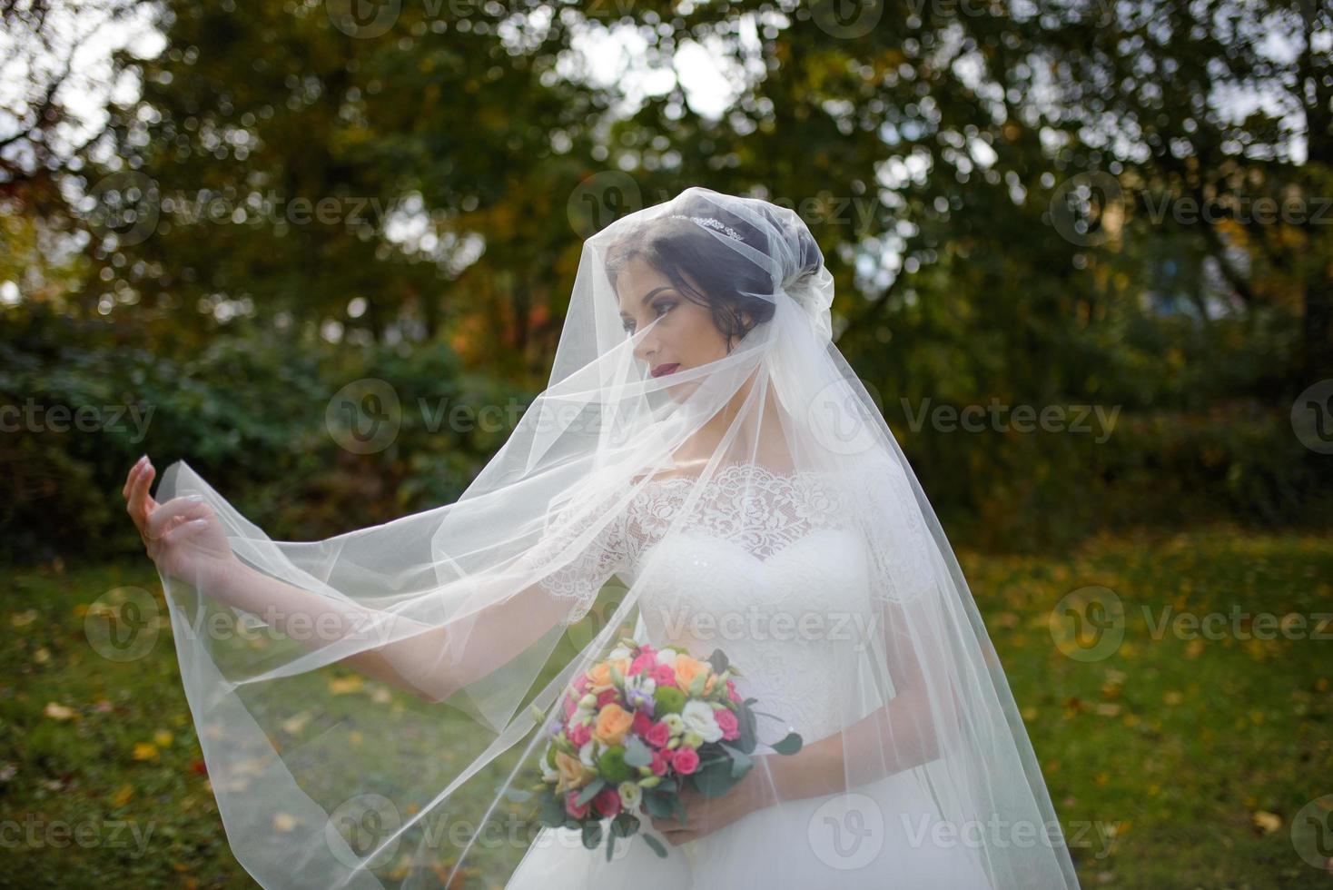 Portrait of a lonely bride on a background of an autumn park. The girl took refuge under a veil with which the wind develops. photo