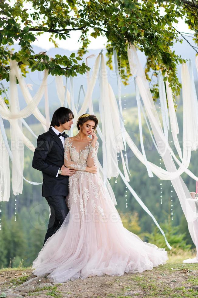 The bride and groom are hugging under an old oak tree. Wedding photo shoot in the mountains. Next to them is prepared decor for the ceremony.