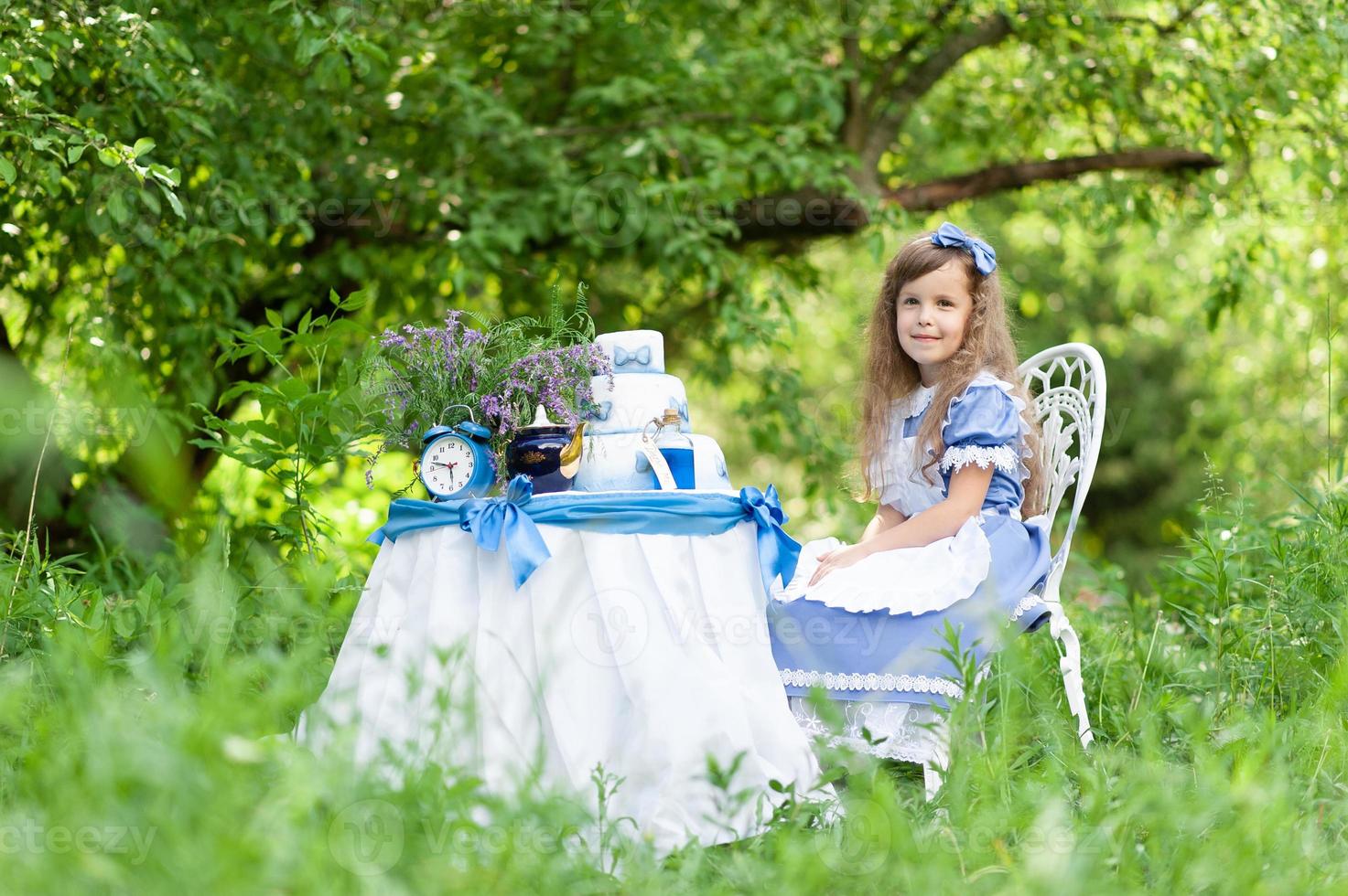 una niña linda con el disfraz de alicia del país de las maravillas organiza una fiesta de té en su mesa mágica. fotografiado en la naturaleza. foto