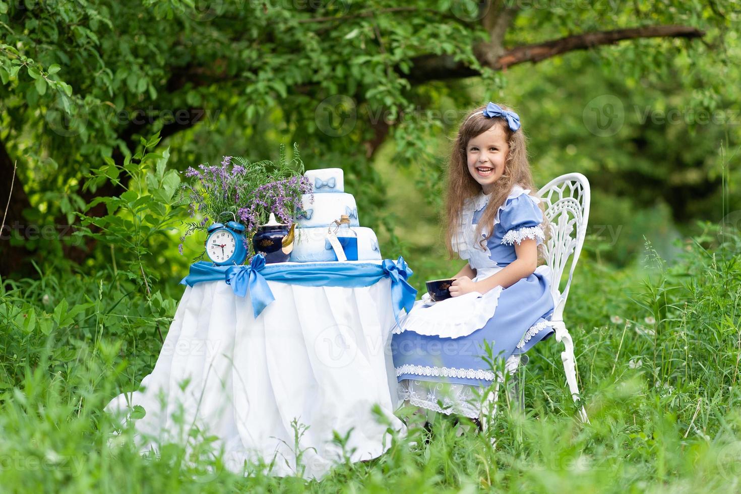 A little cute girl in the costume Alice from Wonderland holds a tea party at her magic table. Photographed in nature. photo