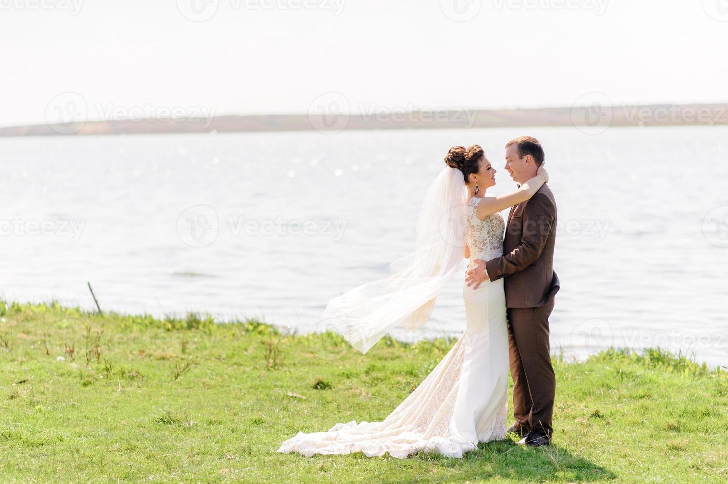 The bride and groom are hugging near the pond in nature. The wind raises a light veil. photo