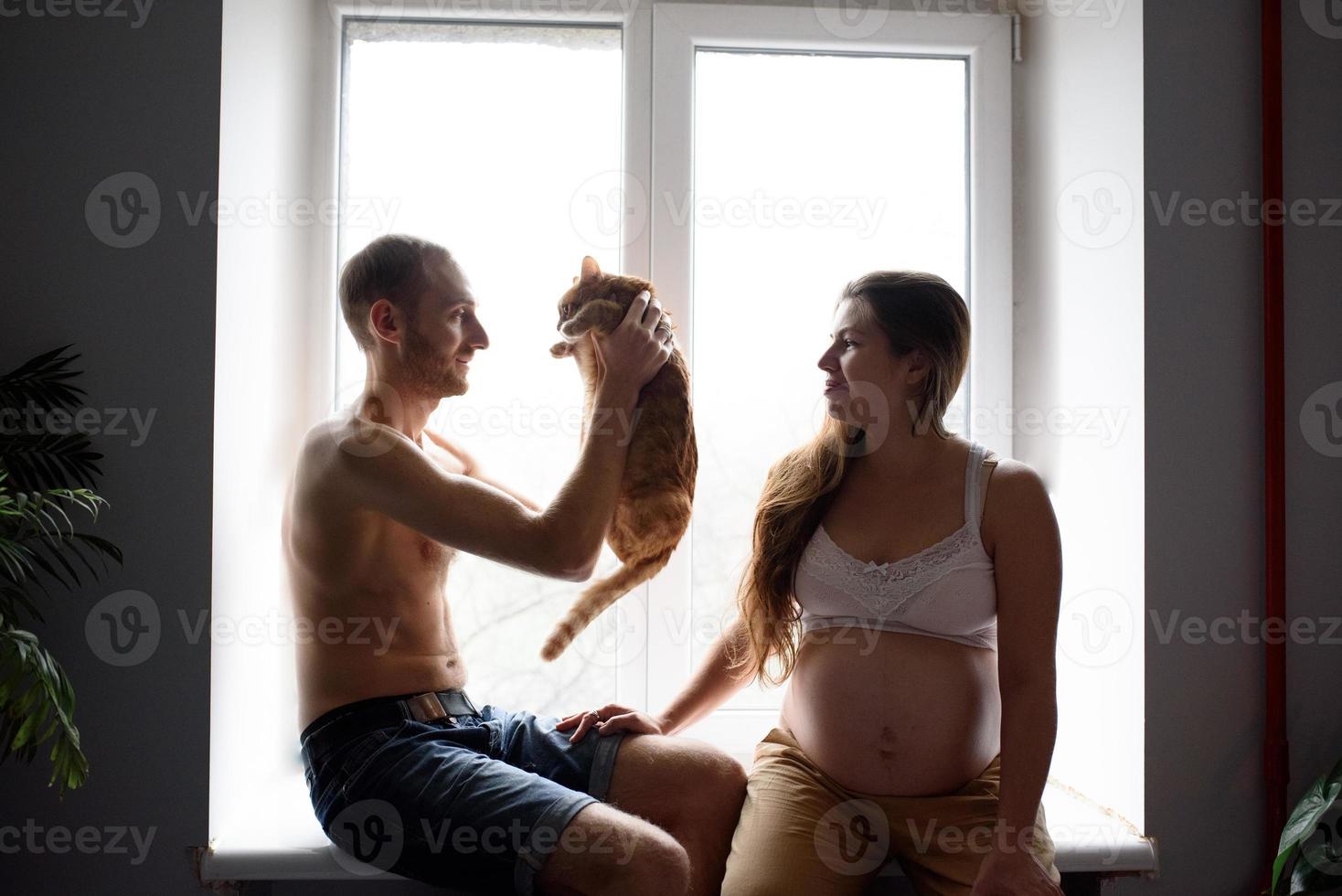 una pareja embarazada enamorada está sentada en el alféizar de la ventana y jugando con su gato. autoaislamiento de una casa durante la cuarentena. foto