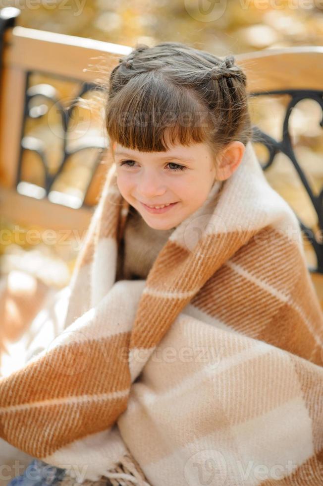 Little cute girl sits on a bench in the autumn park. The girl took shelter in order to warm herself. Autumn time. photo