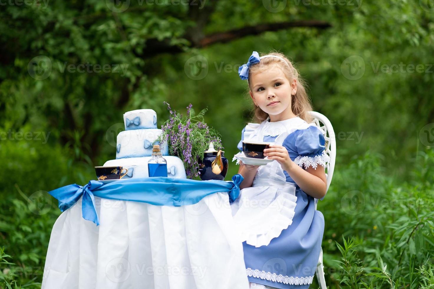 una niña linda con el disfraz de alicia del país de las maravillas organiza una fiesta de té en su mesa mágica. fotografiado en la naturaleza. foto