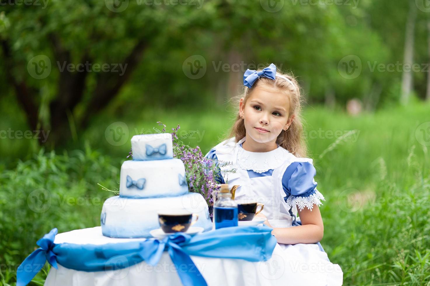 A little cute girl in the costume Alice from Wonderland holds a tea party at her magic table. Photographed in nature. photo