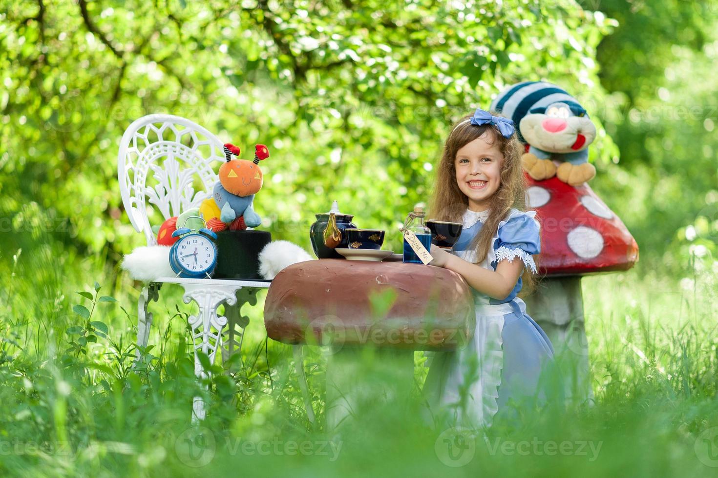 A little cute girl in the costume Alice from Wonderland holds a tea party at her magic table. Photographed in nature. photo