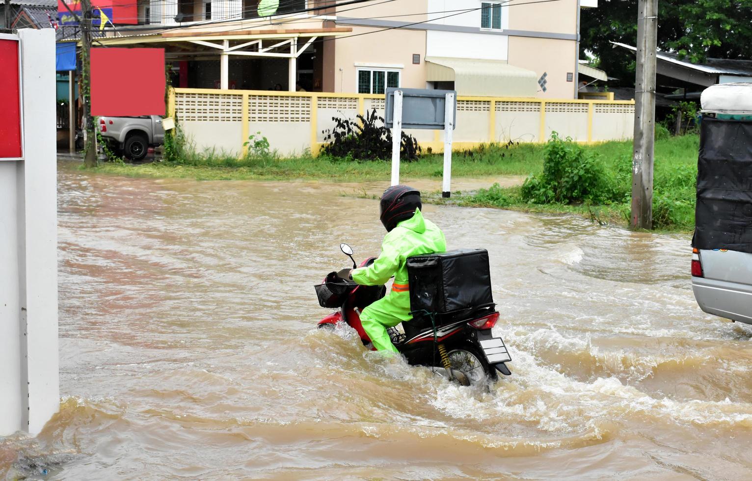 repartidor asiático con uniforme verde, montando y entregando comida al cliente con una caja de comida detrás en la calle inundada, entrega exprés de comida y concepto de compras en línea en la situación de la inundación. foto