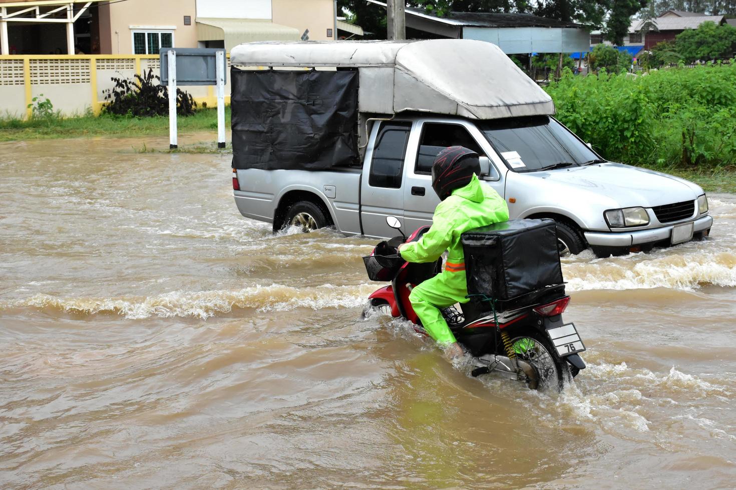 repartidor asiático con uniforme verde, montando y entregando comida al cliente con una caja de comida detrás en la calle inundada, entrega exprés de comida y concepto de compras en línea en la situación de la inundación. foto