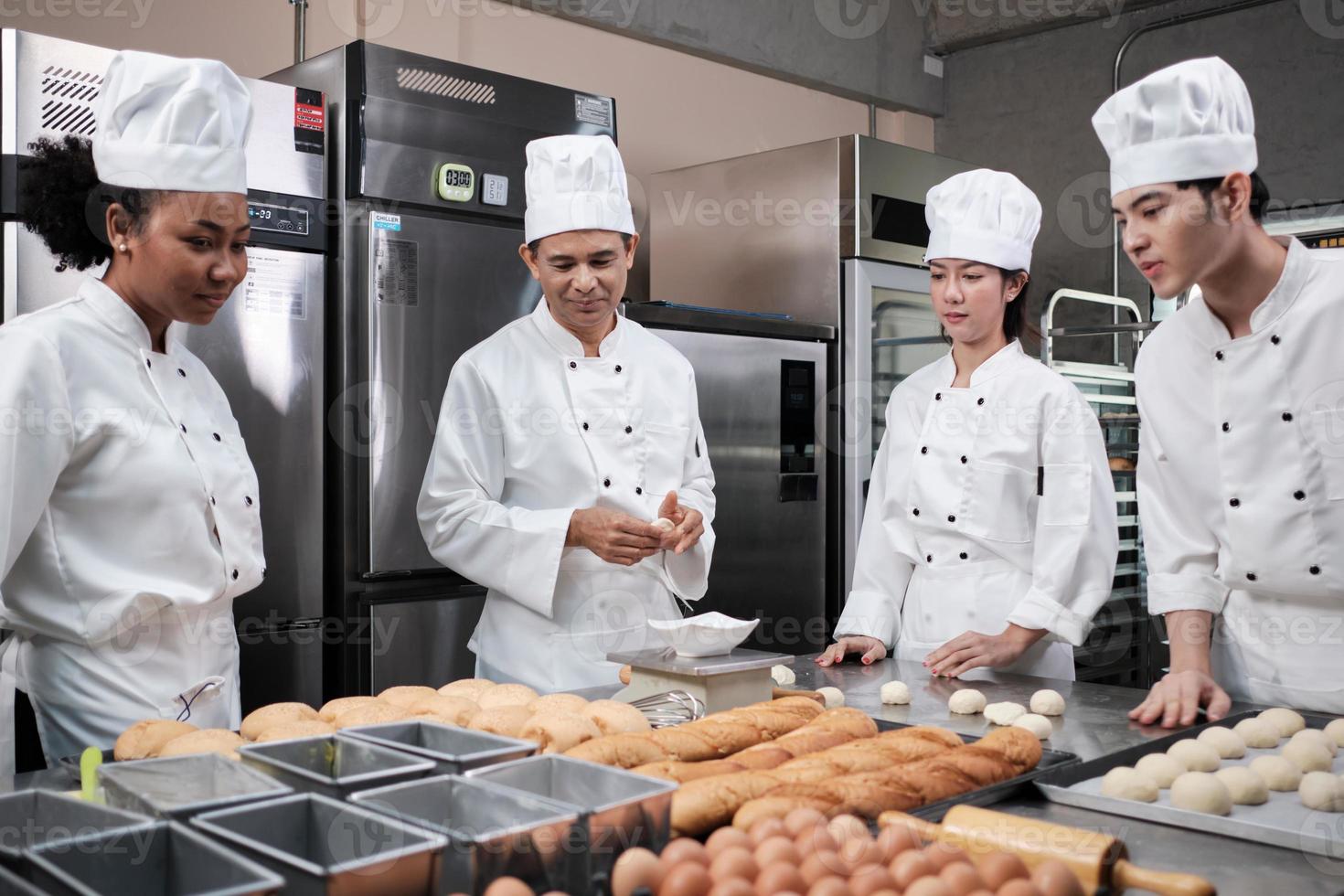 equipo gourmet profesional multirracial, cuatro chefs con uniformes de cocina blancos y delantales amasan masa de pastelería y harina, preparan pan y comida de panadería, hornean en el horno en la cocina del restaurante de acero inoxidable. foto