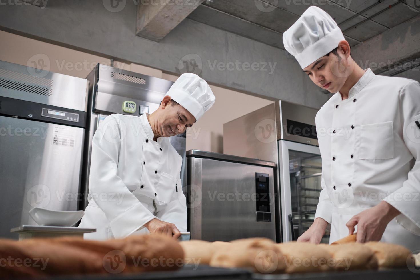 Two professional Asian male chefs in white cook uniforms and aprons are kneading pastry dough and eggs, preparing bread and fresh bakery food, baking in oven at stainless steel kitchen of restaurant. photo