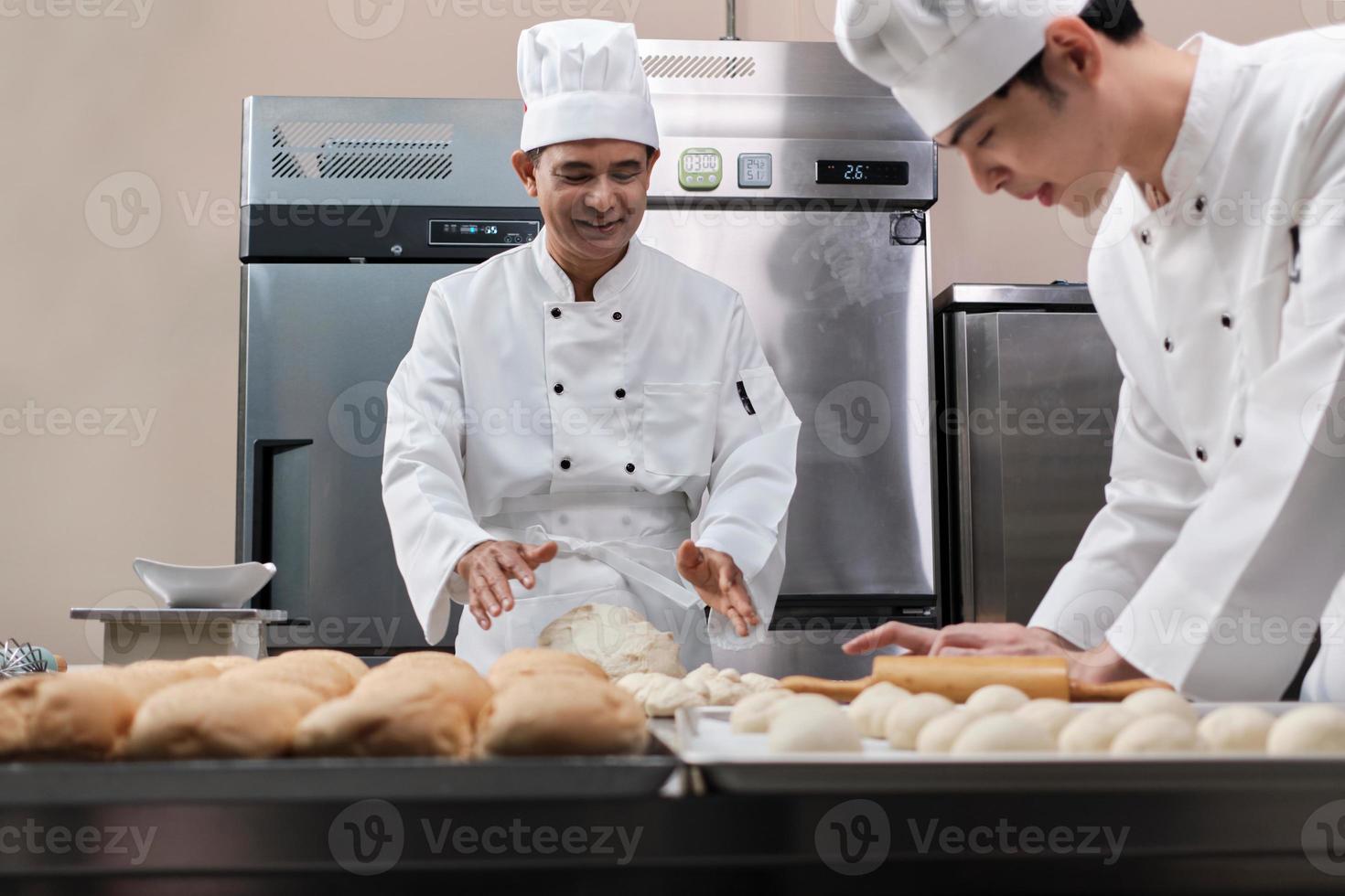 Two professional Asian male chefs in white cook uniforms and aprons are kneading pastry dough and eggs, preparing bread and fresh bakery food, baking in oven at stainless steel kitchen of restaurant. photo