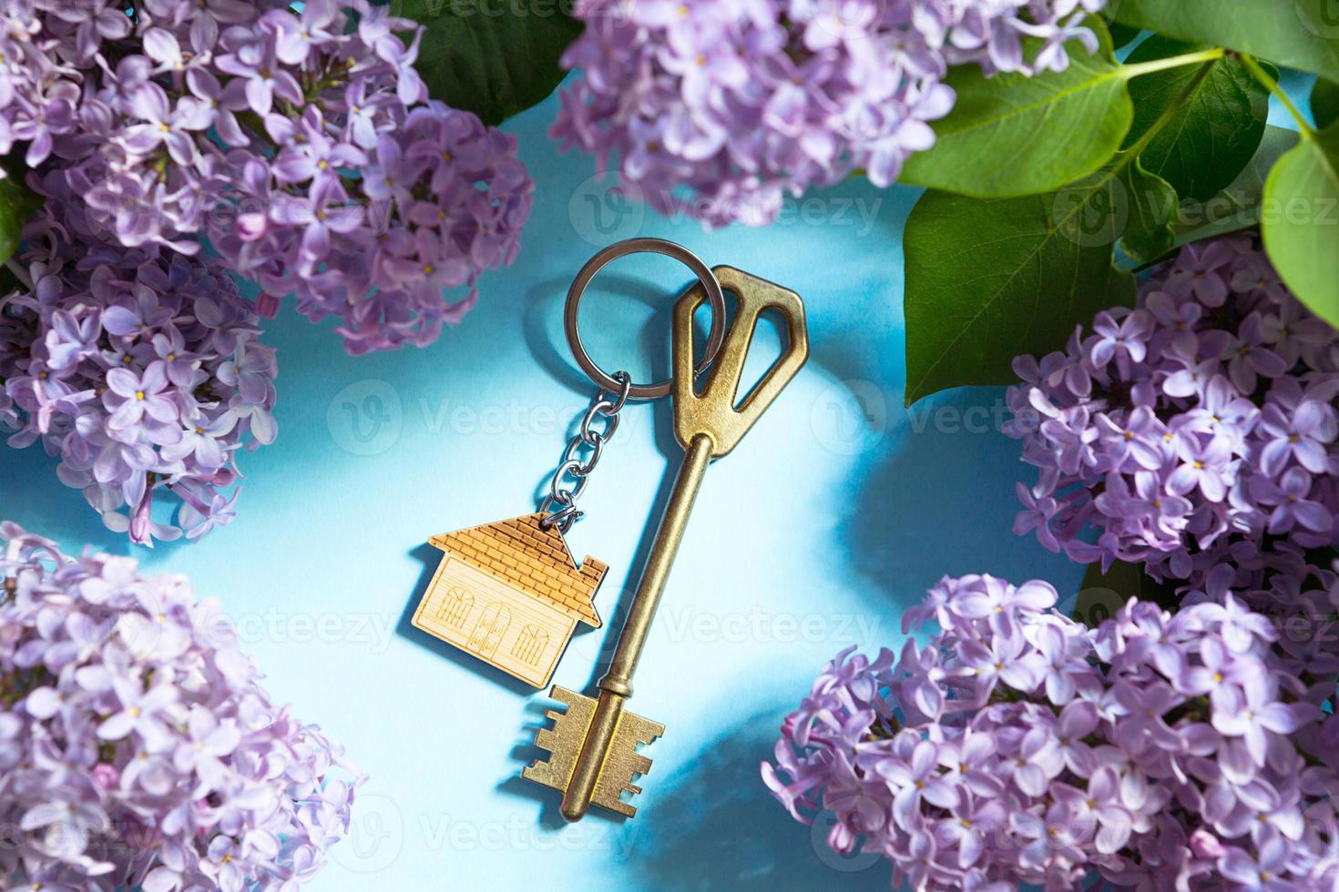 House key with keychain on blue spring background and lilac branches.Summer holiday home, booking cottage in the countryside, moving to new home, mortgage, rent and purchase real estate. Tourism photo