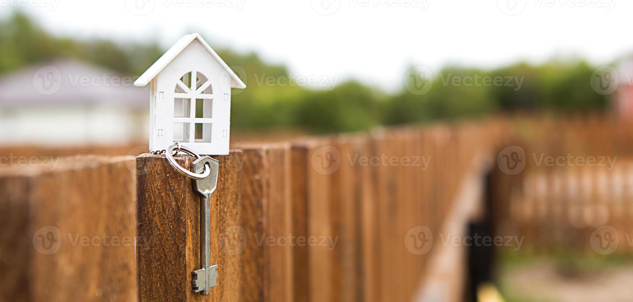 Small figure of white wooden house on fence with key to housing against the background of cottages. Building, design, project, moving to new house, mortgage, rent and purchase real estate. Copy space photo