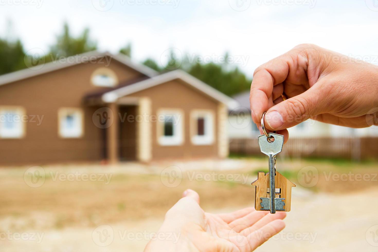 Hand with a key and a wooden key ring-house. Background of fence and cottage. Building, project, moving to a new home, mortgage, rent and purchase real estate. To open the door. Copy space photo
