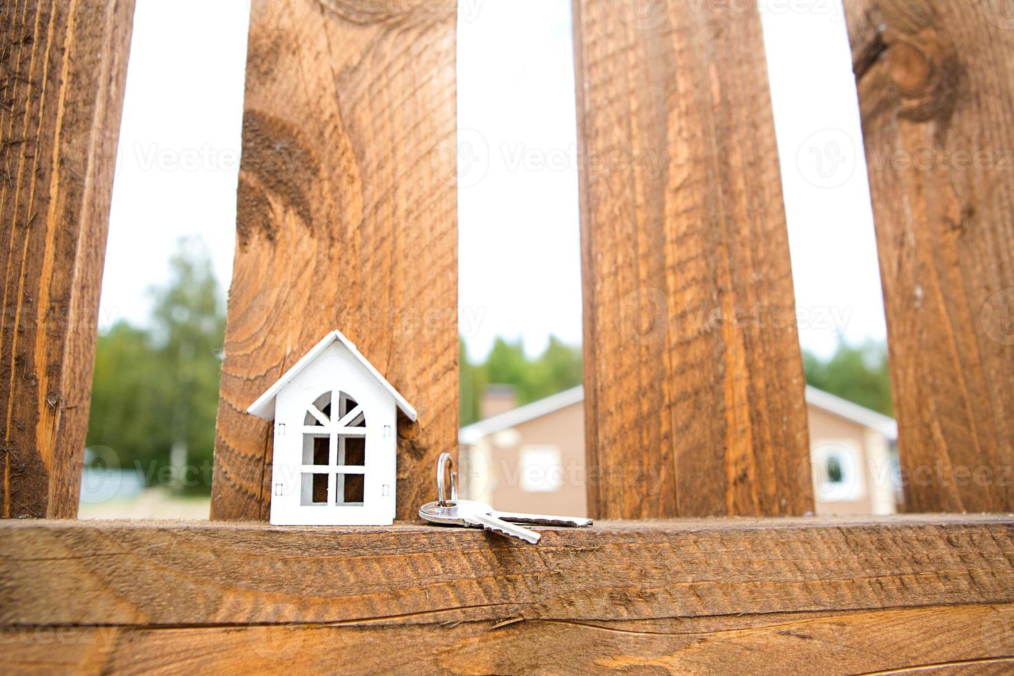 Small figure of white wooden house on fence with key to housing against the background of cottages. Building, design, project, moving to new house, mortgage, rent and purchase real estate. Copy space photo