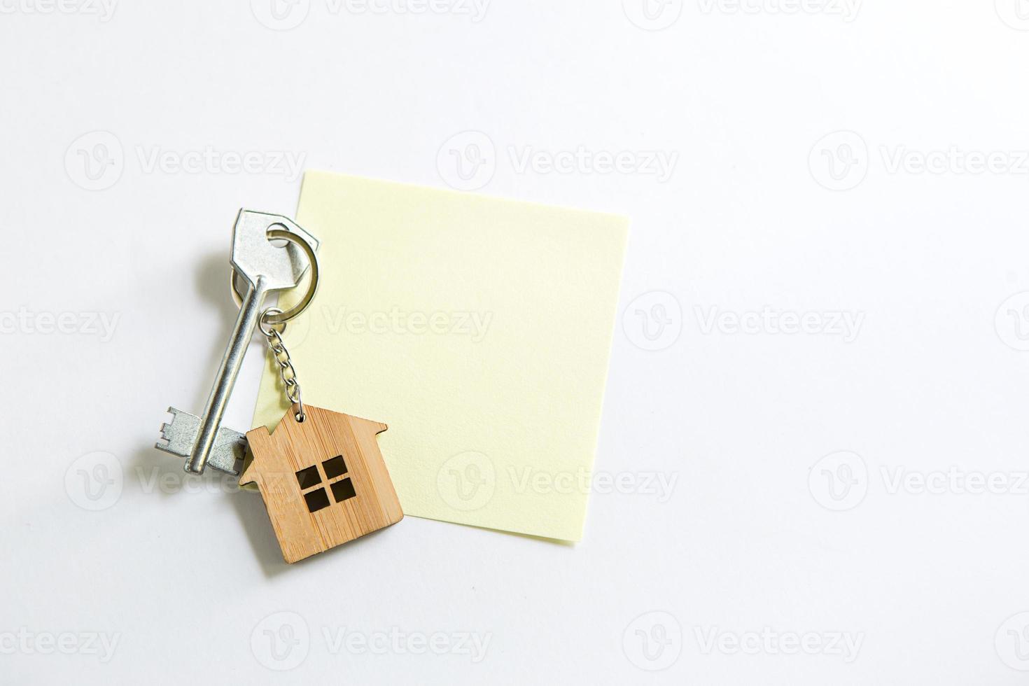 Keychain in the shape of wooden house with key on a white background with a square sheet for notes. Building, design, project, moving to new house, mortgage, rent and purchase real estate. Copy space photo