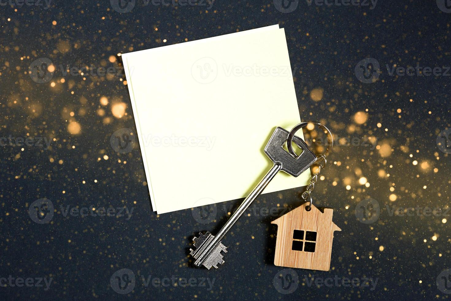 Keychain in the shape of wooden house with key on a black background with a square sheet for notes. Building, design, project, moving to new house, mortgage, rent and purchase real estate. Copy space photo